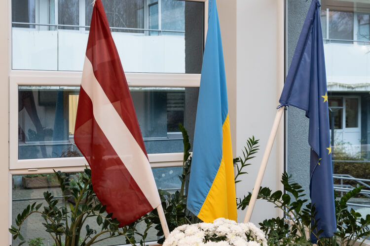 Iekštelpās ir izvietoti trīs karogi: Latvijas karogi kreisajā pusē, Ukrainas centrā un Eiropas Savienības karogi labajā pusē. Ap tiem novietoti augi podos, un priekšā ir vāze ar baltiem ziediem. No lielajiem logiem paveras skats uz āru.