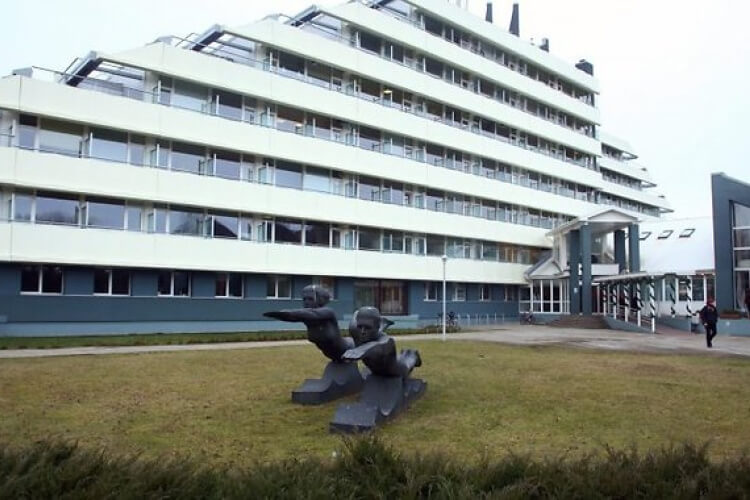 Mūsdienīga, stūraina ēka ar vairākiem stāviem un baltiem balkoniem atrodas aiz zāliena laukuma. Priekšplānā ir tumša divu abstraktu cilvēku figūru statuja. Cilvēks iet netālu no ieejas pa labi.