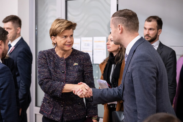 Sieviete tumšā, teksturētā uzvalkā spiež roku vīrietim pelēkā uzvalkā. Tie atrodas iekštelpās ar stikla durvīm fonā. Ap viņiem stāv vairāki citi cilvēki, ģērbušies lietišķā tērpā.