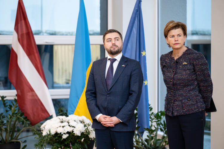Divas personas stāv telpās starp Latvijas, Ukrainas un Eiropas Savienības karogiem. Šķiet, ka viņi atrodas oficiālā pasākumā, ko ieskauj augi podos un baltu ziedu pušķis. Abi ir ģērbušies svinīgā tērpā.
