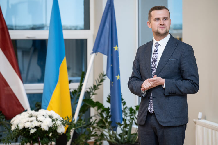 Cilvēks uzvalkā stāv iekštelpās pie Latvijas, Ukrainas un Eiropas karogiem. Šķiet, ka tie atrodas formālā vidē ar augiem un baltiem ziediem tuvumā. Telpas fonā ir lieli logi.