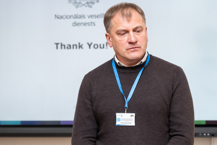Vīrietis brūnā džemperī un zilā štropē stāv pie ekrāna, uz kura redzams uzraksts "Paldies!" Šķiet, ka viņš atrodas konferencē vai prezentācijā.